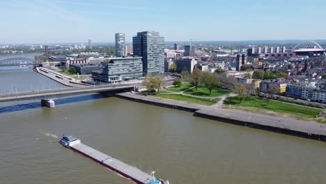Aerial-cityscape-of-industrial-ship-on-urban-river-with-glass-corporate-buildings-in-background,-Cologne