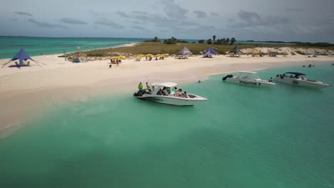 Turquoise-waters-at-Los-Roques-archipelago-with-boats-and-visitors-on-sandy-beach,-sunny-day,-aerial-view