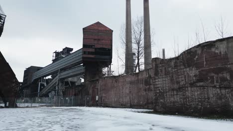 Sitio-Industrial-Desolado,-Cubierto-De-Nieve-Y-Abandonado-Con-Estructuras-Oxidadas,-Un-Edificio-Alto-De-Ladrillo-Y-Dos-Chimeneas-Bajo-Un-Cielo-Nublado