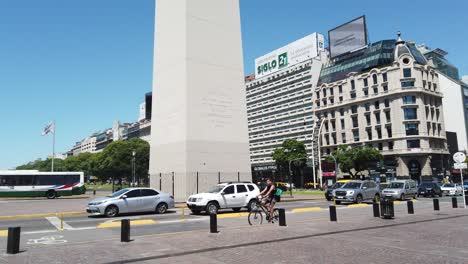 Cars-Drive-Through-Central-Obelisk-Plaza,-Downtown-Center-9-de-Julio-Corrientes-Avenues-with-Morning-Summer-City-Skyline