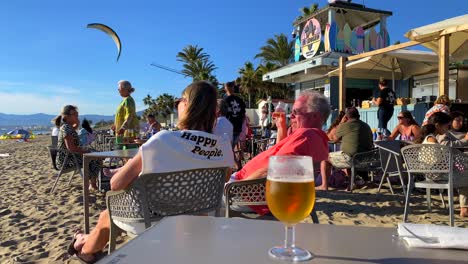 People-sitting,-talking-and-enjoying-life-at-a-beach-restaurant-in-Marbella,-full-beer-glass-on-table,-woman-wearing-a-pullover-with-happy-people-words,-sunny-summer-day-in-Spain,-4K-shot