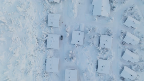 Topview-drone-shot-tracking-a-car-driving-in-middle-of-snowy-cottages-in-Lapland