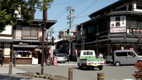 Light-Traffic-Going-Past-Intersection-In-Takayama-Japan,-while-people-walk-and-stroll-on-the-sidewalks,-and-cars-go-along-the-avenues-surrounded-by-businesses-and-restaurants