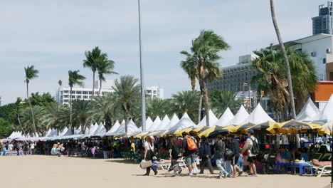 A-group-of-tourists-walking-at-the-right-side-of-the-frame-in-front-of-tents-lined-at-the-coastal-area-of-Pattaya,-in-Chonburi-province-in-Thailand