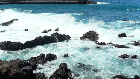 Heavy-ocean-waves-crashing-into-dark-volcanic-rocks-rising-above-surface,-slowmo