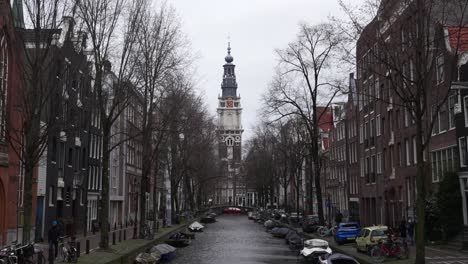 Scenic-static-shot-of-a-canal-in-Amsterdam-with-view-on-a-church,-pedestrians-walking-sidewalk