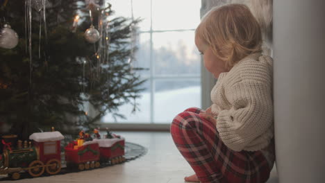 Ein-Kleines-Blondes-Mädchen-Wartet-Auf-Den-Weihnachtsmann,-Während-Es-Zu-Hause-Eine-Spielzeugeisenbahn-Beobachtet