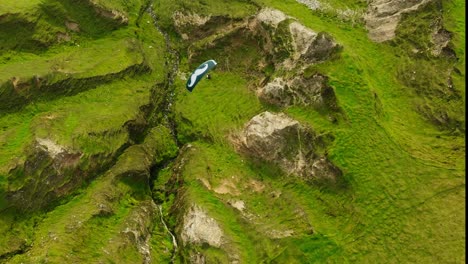 Aerial-birdseye-view-of-a-paraglider-with-a-green-mustached-canopy-sailing-over-bright-green-grass-and-rocky-ravines
