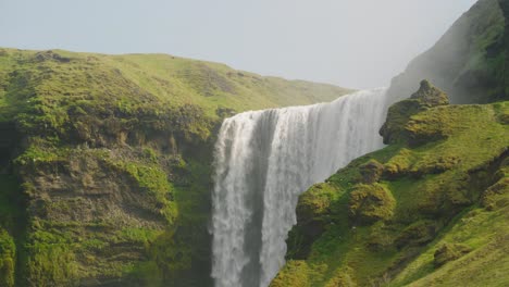Mittlere-Aufnahme-Des-Skogafoss-Wasserfalls-In-Island,-Wunderschöner-Sonniger-Tag-Mit-Vögeln,-Die-über-Den-Moosigen-Klippen-Und-Felsen-Fliegen,-Während-Das-Wasser-Herabstürzt