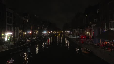 Amsterdam-city-canal-during-night-time,-pedestrians-walking-around-busy-city-area