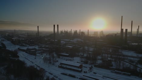 Aerial-view-of-an-industrial-area-blanketed-in-snow,-with-tall,-dark-smokestacks-silhouetted-against-a-vibrant-sky-during-sunset