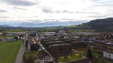 Swiss-town-with-quaint-houses-against-a-backdrop-of-majestic-snow-capped-Alps,-cloudy-skies,-aerial-view