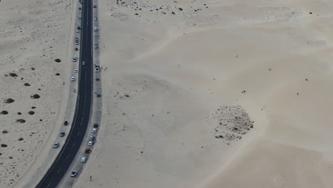 Cars-line-tow-lane-highway-road-in-middle-of-sand-dunes-at-Corralejo-Beach,-aerial-Fuerteventura