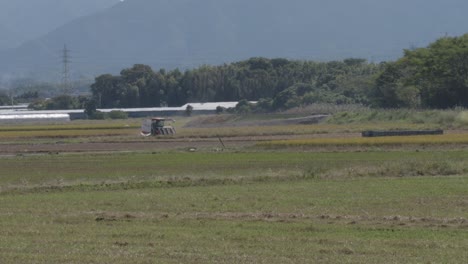 Tractor-Labrando-Tierras-En-Un-Entorno-Rural-Con-Montañas-Al-Fondo,-Día-Soleado-En-Izumi-Japón