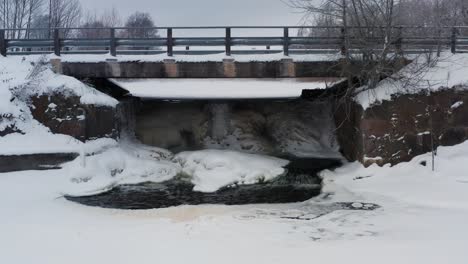Frozen-waterfall-under-small-countryside-bridge,-ice-sheet-cover-on-pond