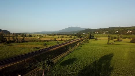 Fly-over-rice-paddy-green-meadow-in-mountain-forest-hills-in-Gilan-near-the-metal-parallel-line-of-railway-train-tourism-transportation-and-traditional-agriculture-concept-harvest-season-cultivation