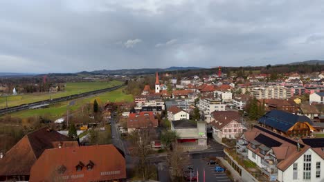 Swiss-town-with-houses,-train-and-countryside,-cloudy-sky,-aerial-view