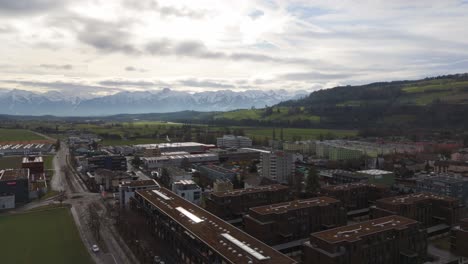 Swiss-town-with-buildings-in-the-foreground-and-snow-capped-mountains-in-the-distance,-cloudy-skies-above,-aerial-view