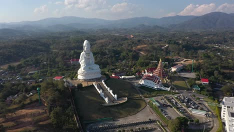 Backwards-aerial-drone-of-Wat-Huay-Pla-Kang-giant-white-big-statue-and-pagoda-temple-with-mountains-and-landspace-in-Chiang-Rai,-Thailand