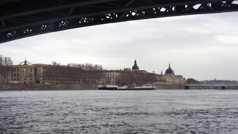 Cargo-Ship-Glides-Along-the-Serene-Rhone-River,-Beneath-Lyon's-Arched-Steel-Bridge,-Amid-Old-European-Cityscapes