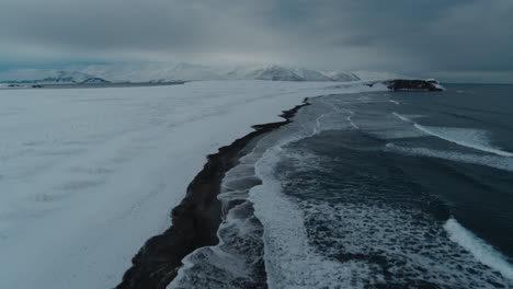 Snow-covered-beach-with-black-sand-and-dark-water-in-Iceland-during-winter-shot-from-a-drone