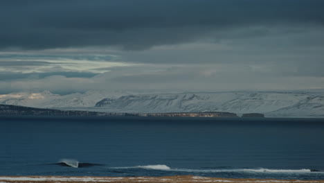 Empty-arctic-wave-from-a-distance-landing-on-shallow-reef-with-snow-mountains-surrounding