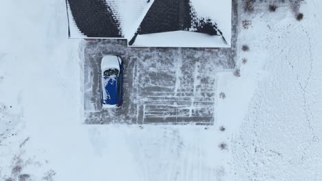 Overhead-drone-shot-of-a-blue-car-in-a-snow-covered-driveway