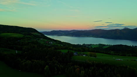 Luftaufnahme-Eines-Sees,-Umgeben-Von-Bergen-Und-Einigen-Kleinen-Häusern-Am-Farbenfrohen-Sonnenuntergangshimmel