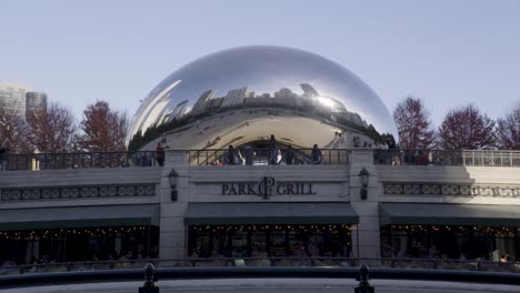 Cloud-Gate-sculpture-reflecting-Chicago-skyline,-with-people-and-Park-Grill-restaurant