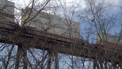 Intermodal-Freight-Train-Crosses-the-Pope-Lick-Trestle-in-Louisville,-KY,-Behind-some-Bare-Trees-on-a-Cloudy-Morning