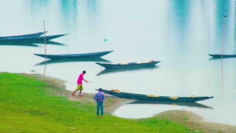 Two-people-walking-by-moored-wooden-boats-on-a-tranquil-riverbank-in-Bangladesh