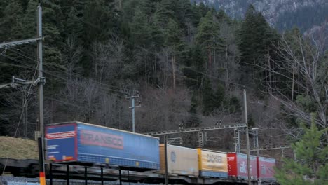 Freight-train-passing-through-a-forested-mountain-region-at-dusk,-cargo-containers-in-view