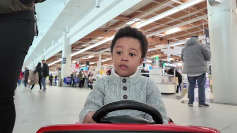 Schöner-Dreijähriger-Exotischer-Schwarzer-Junge,-Der-Ein-Elektrisches-Spielzeugauto-In-Einem-Einkaufszentrum-Fährt,-Gefolgt-Von-Seiner-Mutter