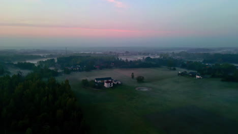 Panorama-Luftaufnahme-Einer-Riesigen-Grünen-Wiese-Mit-Einigen-Häusern,-Umgeben-Von-Natur,-Bäumen-Und-Nebel