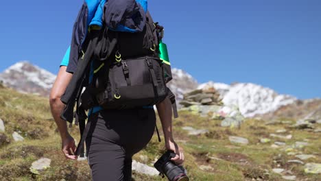 Slow-motion-man-with-backpack-and-camera-climbs-up-toward-mountain-peaks-on-sunny-day