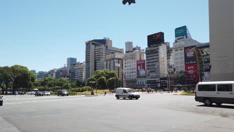 Car-traffic-drives-along-9-de-Julio-Avenue-in-the-city-center-in-summer,-next-to-the-obelisk-and-commercial-stores