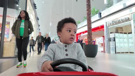 Sweet-and-amused-3-year-old-balck-kid-riding-an-electric-toy-car-inside-a-mall-followed-by-his-mother