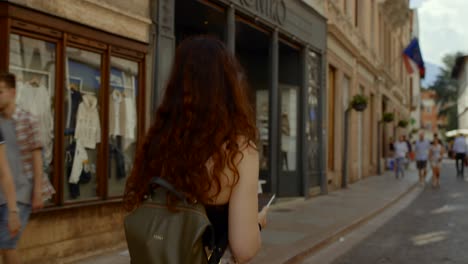 Tracking-shot-of-young-woman-with-wavy-hair-walking-down-a-street-with-shops-in-Verona,-Italy