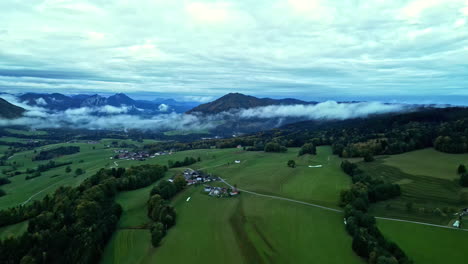 Wunderschöne-Luftaufnahme-Einer-Riesigen-Grünen-Wiese-Mit-Einigen-Häusern,-Umgeben-Von-Bergen,-Natur-Und-Niedrigen-Wolken