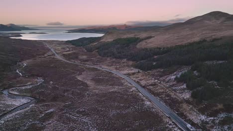 Winding-road-through-rugged-terrain-at-dusk,-light-snow-cover,-serene-highland-landscape,-aerial-view