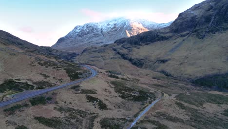 Glencoe-with-snow-capped-mountains-and-winding-road-at-dusk,-serene-highland-landscape,-aerial-view