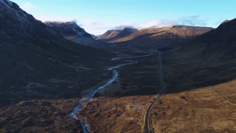 Glencoe-valley-with-winding-river-and-road-amidst-rugged-mountains,-daylight,-aerial-view