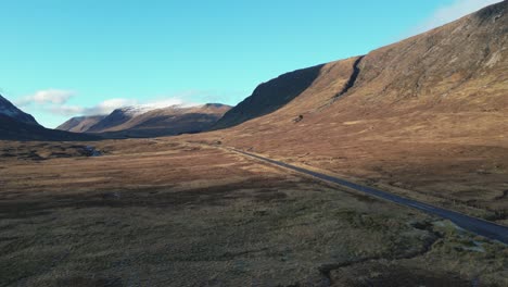 Remote-valley-road-winding-through-Glencoe-with-towering-mountains,-clear-skies,-Scotland,-aerial-view