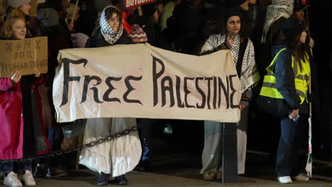 Two-women-wearing-Keffiyeh-scarves-hold-a-white-banner-that-reads,-“Free-Palestine”-during-a-nighttime-protest-outside-the-Houses-of-Parliament