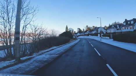 Winding-road-with-snow-patches-on-the-sides-leading-through-a-serene-Scottish-landscape,-clear-blue-skies,-winter,-car-POV