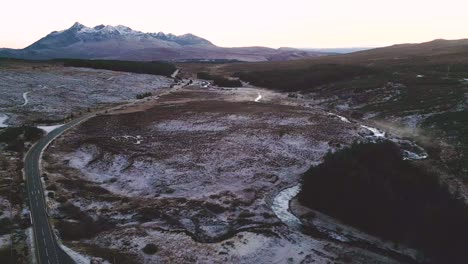 Winding-road-through-snow-dusted-landscape-with-mountain-backdrop-at-dusk,-aerial-view