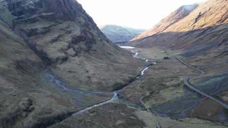 Glencoe-Valley-with-meandering-river-and-winding-road-amidst-rugged-mountains,-early-morning-light,-aerial-view
