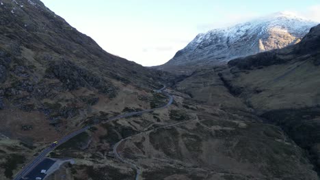 Winding-road-through-the-rugged-terrain-of-Skye-with-snow-capped-mountains-in-the-backdrop,-dusk-light,-aerial-view