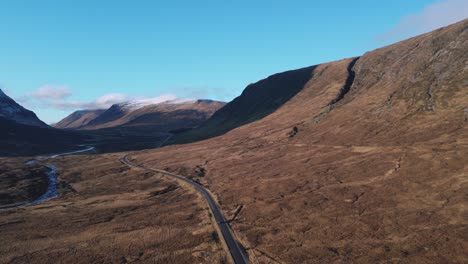 Meandering-road-in-Glencoe-valley-with-rugged-mountains-and-clear-skies,-aerial-view