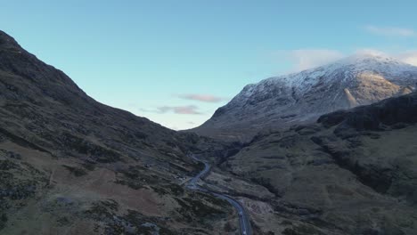 Winding-road-through-the-snow-capped-Glencoe-mountains-at-dusk,-serene-and-majestic,-aerial-view